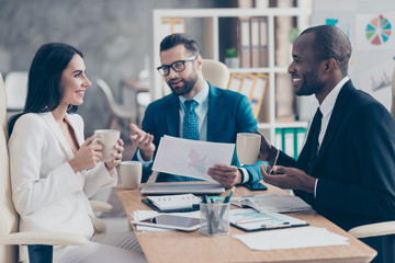 Three professional, successful, stylish, elegant employees having break time during conference, speaking, drinking tea holding mugs with coffee in hands, sitting in work place, station