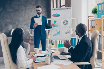 Stylish, bearded, handsome employee holding tablet in hand, pointing on diagrams on the white board, coach making consultation, presenting results on blurred background in work place, station