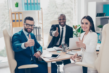 Three attractive, stylish, smart, confident businesspeople, agents in elegant classic suits sitting on workstation, workplace, showing thumb up symbol, looking at camera