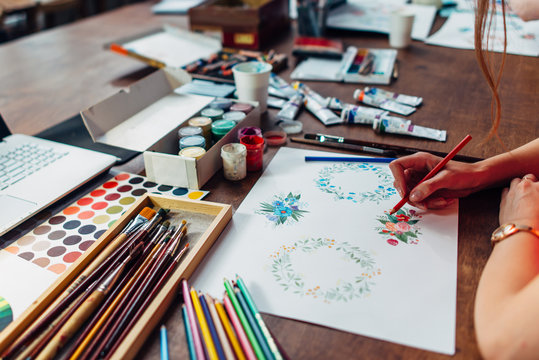Close-up of female designer drawing floral compositions with crayons sitting at workplace surrounded with paint, gouache, brushes and other art supplies