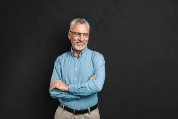 Obraz premium Portrait of caucasian male pensioner 60s with grey hair and beard smiling and looking aside while standing with arms folded, isolated over black background