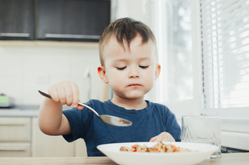 beautiful cute baby eats rice with a spoon in the kitchen, very fun