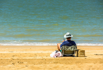 Senior man reading a newspaper on the beach at the water's edge.