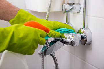 Man in rubber protective gloves cleaning and polish shower faucet. Shiny surface of water tap, hands with sponge and bottle of cleaning agent. White tiles of bathroom