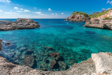Wide angle view of Crystalline turquoise waters of Cala s'Almunia beach from cap des moro. Small boat sailing and People swimming. Located in Santanyi, Majorca, Balearic Islands, Spain.