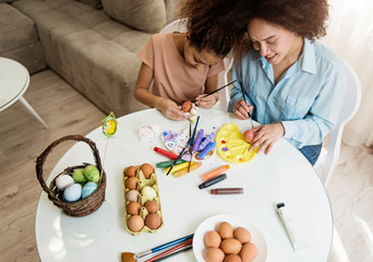 Beautiful African American woman and her daughter coloring Easter eggs at the table  