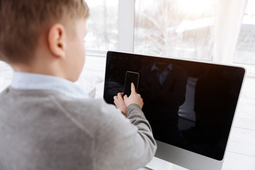 Digital device. Smart cute positive boy sitting in front of the digital screen and holding a smartphone while using this device