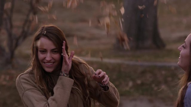 Teenage girl thowing leaves on twin sister at dusk in park during Fall.