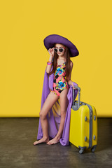 Attractive girl teenager 11 years in swimsuit, hat, sunglasses, suitcase. Stunning girl model in a colorful bikini and pareo posing on a bright yellow background. Studio shot. Concept summer, travel.