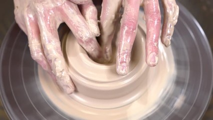 Muddy potter hands are shaping a clayware from the lump of clay on the spinning potter's wheel.