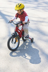 Little child boy cycling on bicycle in green park outdoor in spring. A child is riding a children's bike with support training wheels wearing safety helmet
