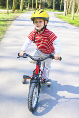 Fototapeta premium Little child boy cycling on bicycle in green park outdoor in spring. A child is riding a children's bike with support training wheels wearing safety helmet 