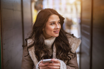 Naklejka premium Close up portrait view of attractive beautiful young smiling girl in sweater and jacket holding a mobile and looking far away while standing in the middle of two wooden walls outside.