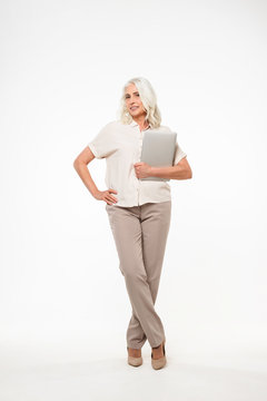 Full Length Portrait Of Mature Adult Woman 60s With Gray Hair Smiling And Holding Silver Laptop In Hand, Isolated Over White Background