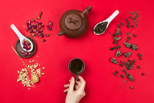 Flatlay frame arrangement with Chinese green tea leaves, rose buds, jasmine flowers, tea pot and woman's hands holding tea cup. Red background