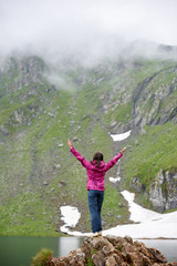 Woman is standing with her back on a rock with her arms wide open and looks up against background of mighty mountains and the lake at the foot, Transylvania, Romania.