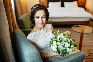Elegant charming young brunette bride in the hotel room  is sitting on sofa in a wedding dress with...