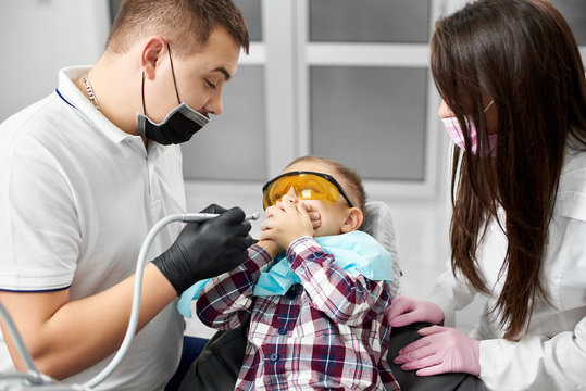 A small boy in a dental chair closes his mouth with his hands, fearing a dentist who has a drill in his hand. Modern dental clinic
