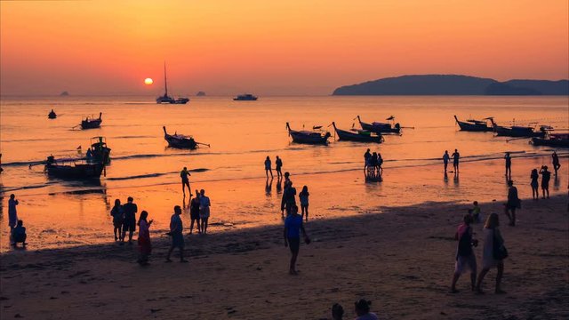 Time lapse of the Thai traditional beach with a lot of people silhouettes, moored Thai long-tail boats and yachts at sunset.