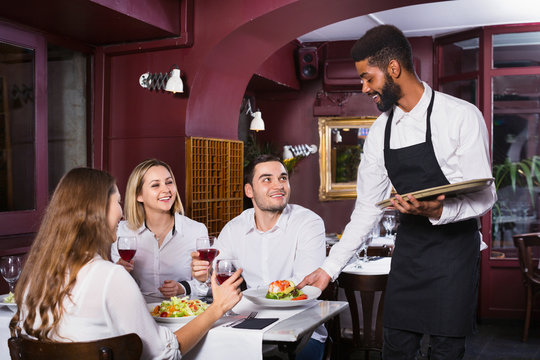 Young cheerful waiter taking care of adults