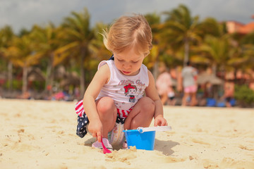 Cute Adorable Two Years Old Toddler Girl with Blonde Hair and Big Blue Eyes in Summer Dress with American Flag Print is Playing with Sand at the Beach, Stormy, in Cancun Resort in Mexico, March 2018