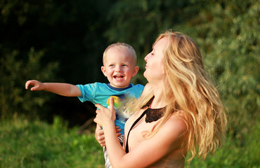 Fototapeta premium Young blonde mother in playing with her child in the green lawn. Happy fun summer photo. Smiling laughing boy with his mom. Sunny day outdoors in nature. Warm hug and embrace.