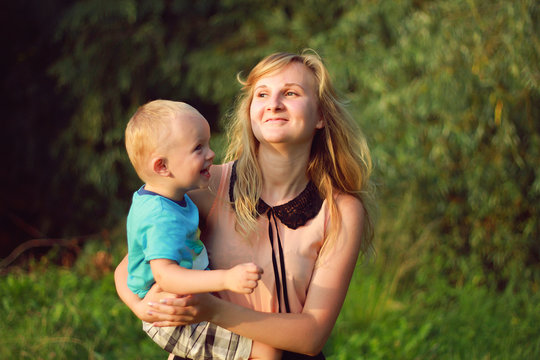 Young blonde mother in playing with her child in the green lawn. Happy fun summer photo. Smiling laughing boy with his mom. Sunny day outdoors in nature. Warm hug and embrace.