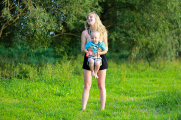 Mother with blonde hair is hugging her child in the green lawn. Happy fun summer photo. Smiling laughing boy with his mom. Sunny day outdoors in nature. Kid and mom are hugging. Warm embrace.