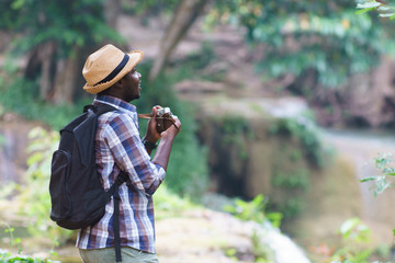 African Man Traveler with backpack standing and holding camera with waterfall background.