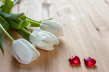 three white tulips and two red hearts on a light wooden background