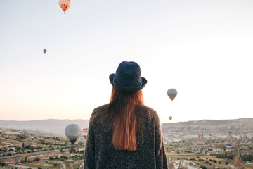 A tourist girl in a hat admires hot air balloons flying in the sky over Cappadocia in Turkey. Impressive sight.