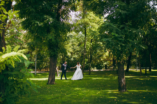 Beautiful wedding photosession. Handsome groom in blue formal suit with bouquet and his elegant bride in white dress and veil with beautiful hairdress on a walk in the big green park on a sunny day