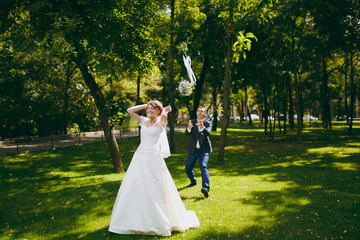 Beautiful wedding photosession. Handsome groom in blue formal suit catches a bouquet that elegant bride throws in dress and veil with beautiful hairdress on a walk in the big green park on sunny day