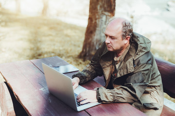 Adult male gamekeeper is sitting outdoor at a wooden table with his gadgets and using the netbook;...