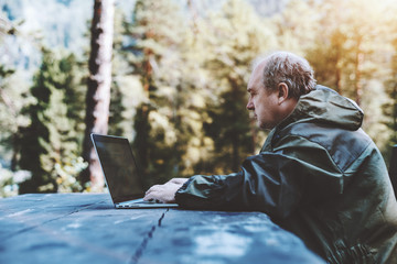 Side view of an aged male forester in dark-green coverall making a report on his laptop while...