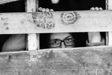 Locked up guilty girl in glasses emotional portrait. Woman slavery. Werid strange unusual person looking through slit in boarded up window in old wooden abandoned ghost house. Female eyes expression.