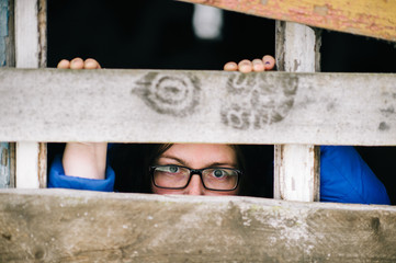 Locked up guilty girl in glasses emotional portrait. Woman slavery. Werid strange unusual person looking through slit in boarded up window in old wooden abandoned ghost house. Female eyes expression.