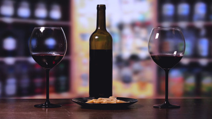 On the table there is a crystal glass, a bottle of red wine, cheese in a black plate, a background of a bar counter.