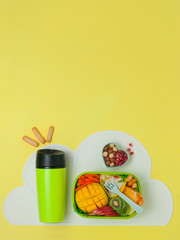 Offene Lunchbox mit Reis, frischem Obst und Gemüse und Thermobecher auf gelbem Hintergrund