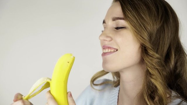 Seductive fair haired young woman in gray peeling a banana and eating it. She is looking at you. Locked down real time close up shot