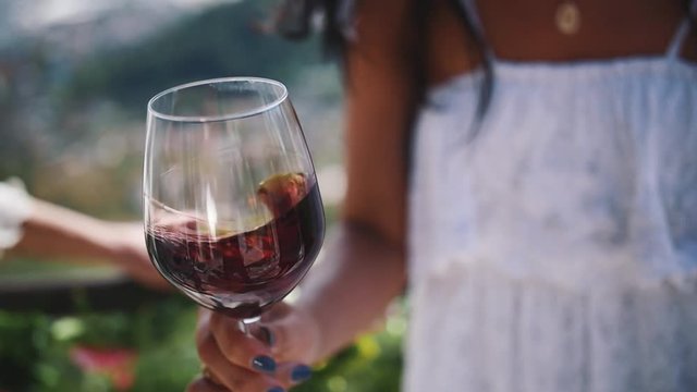Hand of young unrecognizable woman in white dress shakes glass of red wine outdoors on hot summer sunny day