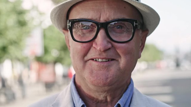 Close up shot of elderly man in hat and glasses standing in pedestrian street on warm day, smiling and looking at camera, tilt up