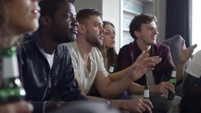 Multi-ethnic group of friends sitting on couch in living room and having beer while watching soccer game on TV and getting disappointed about team failure
