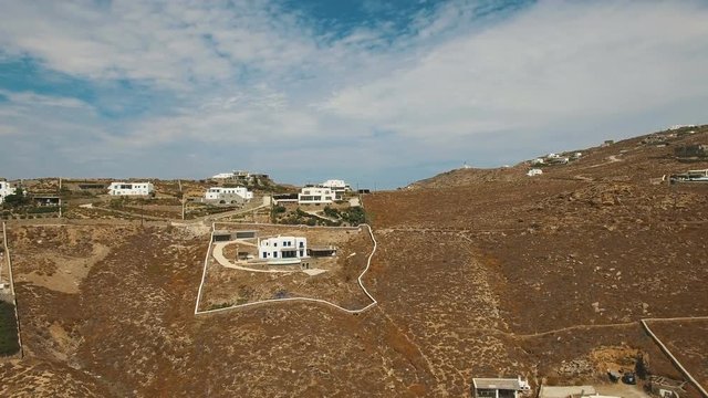 Black car moving up the road to the hill with luxury white villas on top. Greek Cyclades with dry land, brown stones and arid bushes. Faros Armenistis, Mykonos island, Greece, Aerial.
