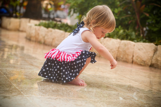 Cute Adorable Two Years Old Toddler Girl with Blonde Hair and Big Blue Eyes in Summer Dress with American Flag Print  is Dancing at the Rain, by the Pool Area in Cancun Resort in Mexico, March 2018