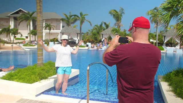 A husband takes a picture of his wife with a cell phone. They are at the pool at a tropical resort.