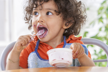 Toddler sitting in highchair and eating greek yogurt. Baby learning to eat and has yogurt on face...