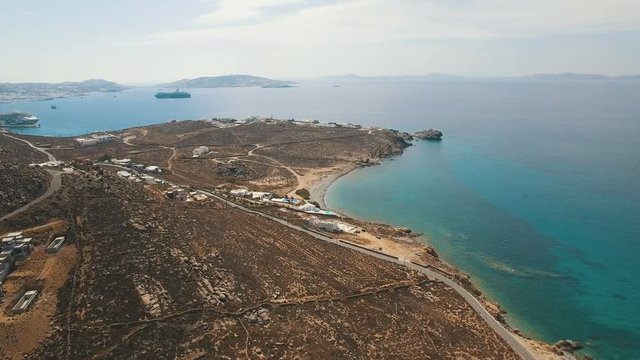 Flying above dry land with brown stones, boulders, arid bushes and dusty roads towards the Aegean sea coast line with beach and white villas. Cyclades. Faros Armenistis, Mykonos island, Greece. Aerial