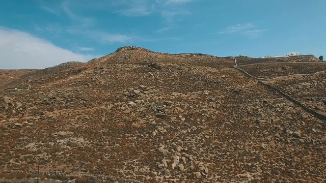 Flying up dry brown ground hill with stones, boulders, arid bushes, dusty roads and historical ruins. Typical scenic landscape of the Greek Cyclades. Faros Armenistis, Mykonos island, Greece. Aerial.