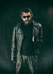 A disheveled and intoxicated man wearing a black leather jacket, sunglasses and holding a bottle of beer.